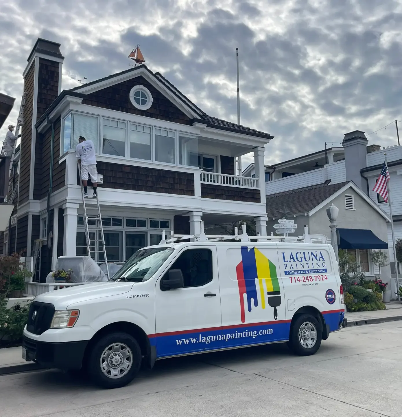 Two painters on ladders painting the exterior of a two-story house while a Laguna Painting van is parked in front.