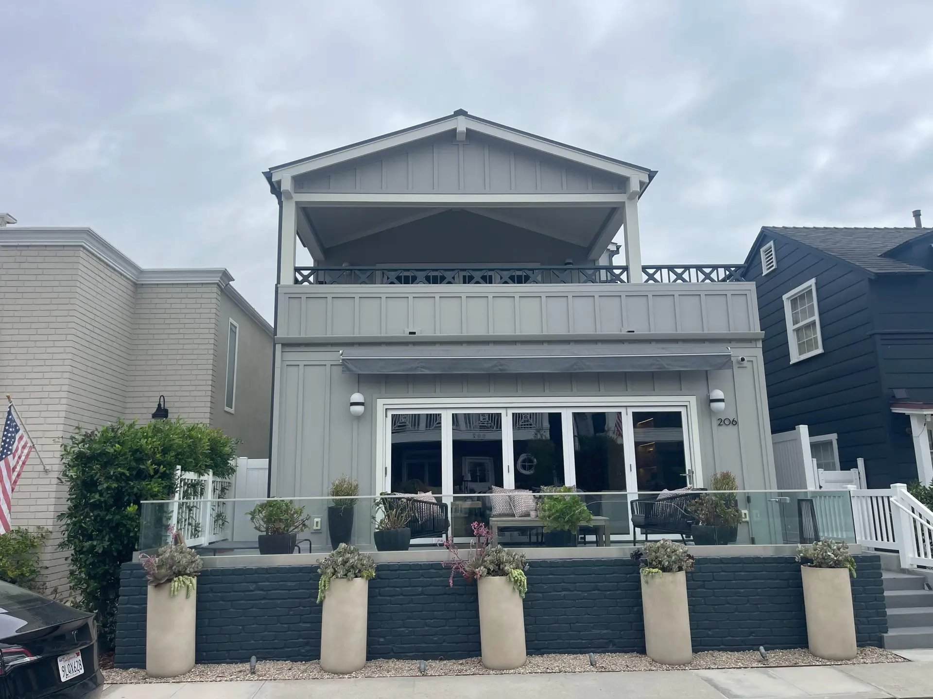 Two-story gray house with a covered upper deck, glass railing, and a front patio with seating; tall planters line the base.