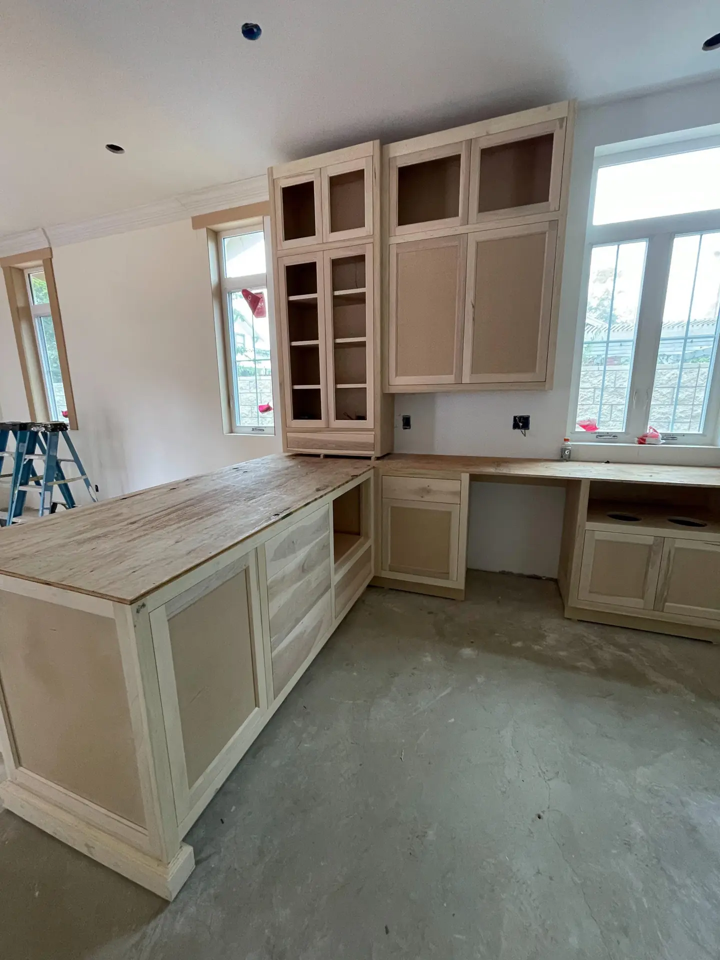 Unders construction kitchen with light wood cabinets, an island, and upper cabinets; tall glass-front cabinet and windows in a bright room.