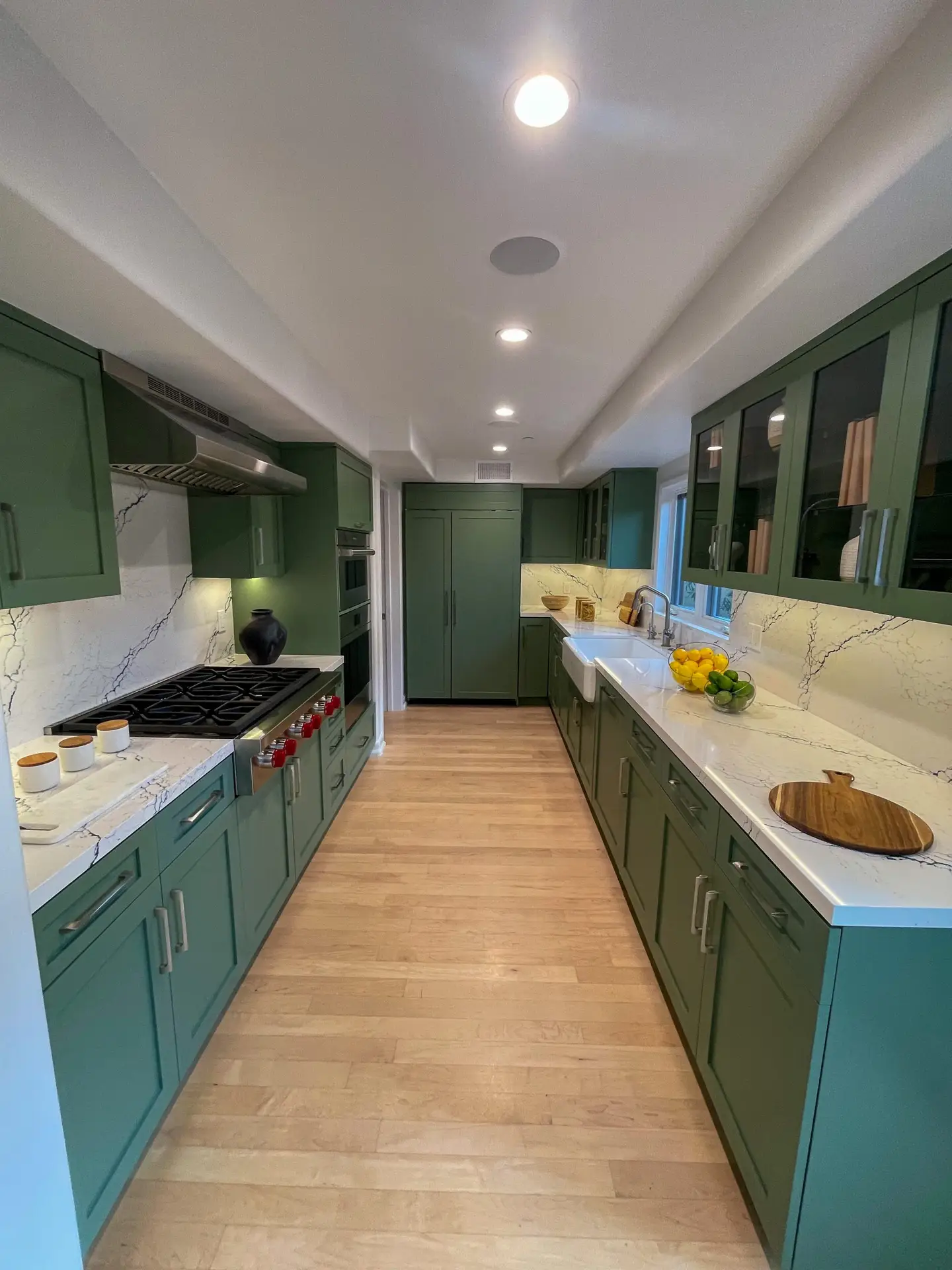 Long green-garden kitchen galley with marble countertops, stainless range, and an islandless layout; fruit bowl near sink and wooden cutting board on counter.