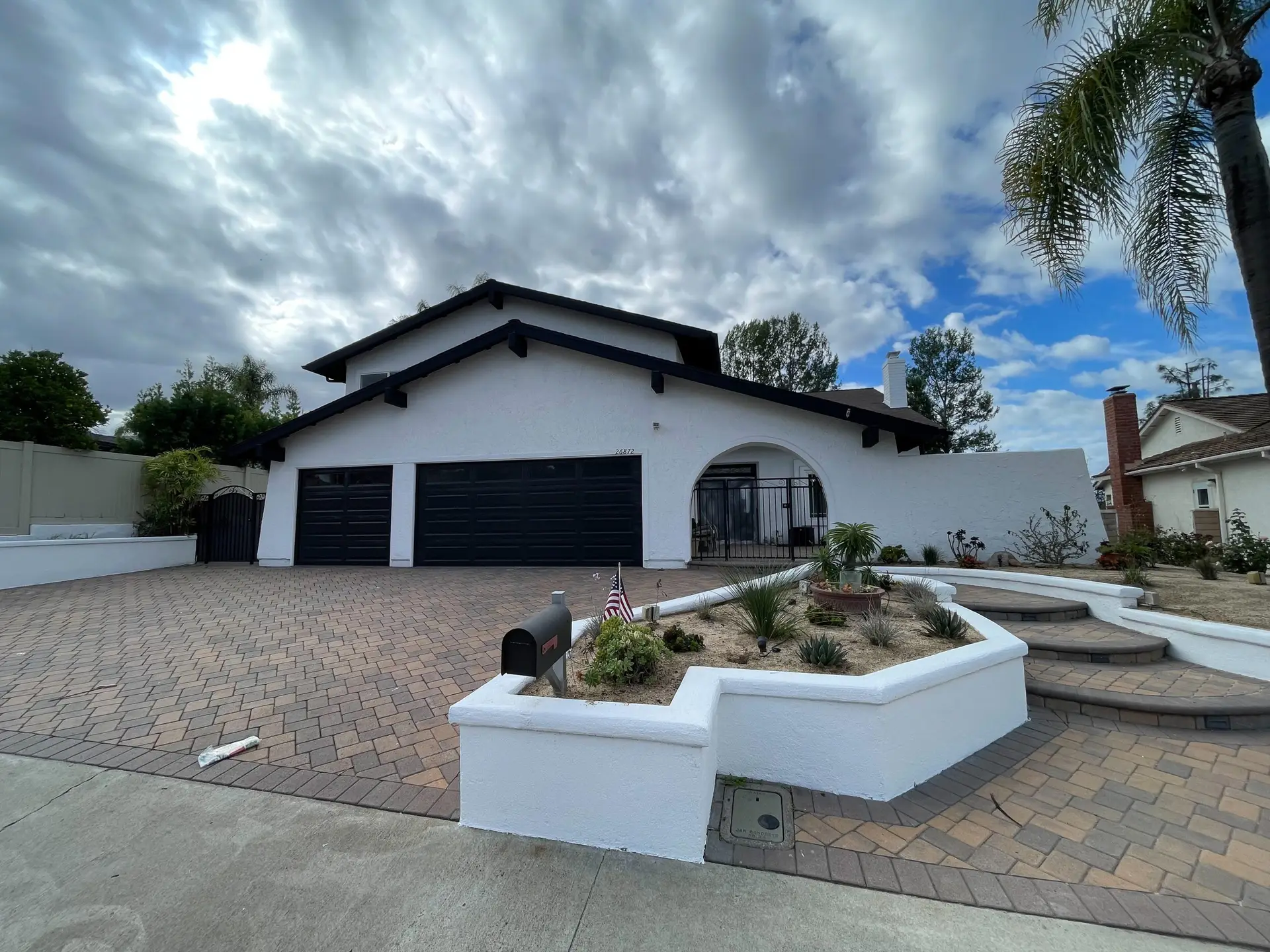 White stucco house with a 3-car black garage and curved entry arch, set behind a low white planter and desert landscaping.