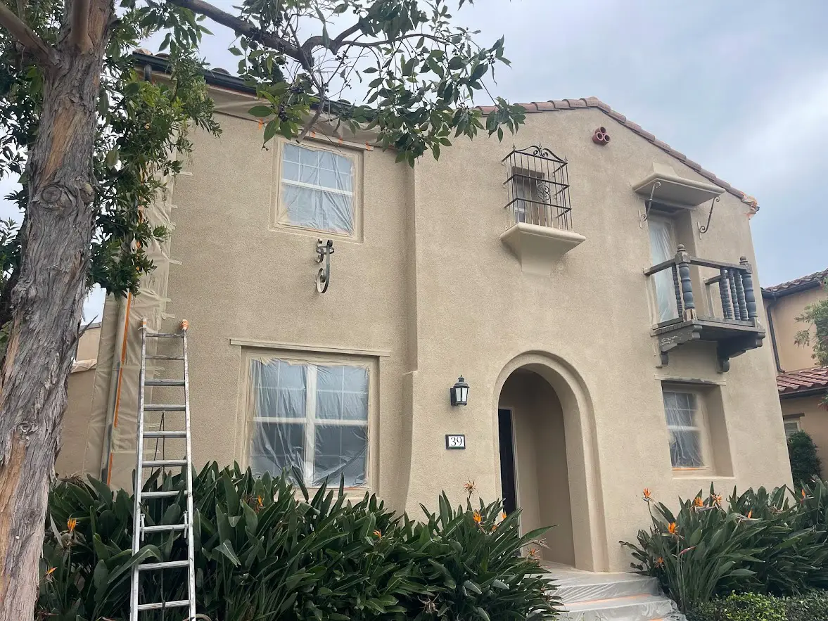 Two-story beige stucco house with arched entry, ladder leaning against the front wall, and plastic-covered windows amid landscaping.