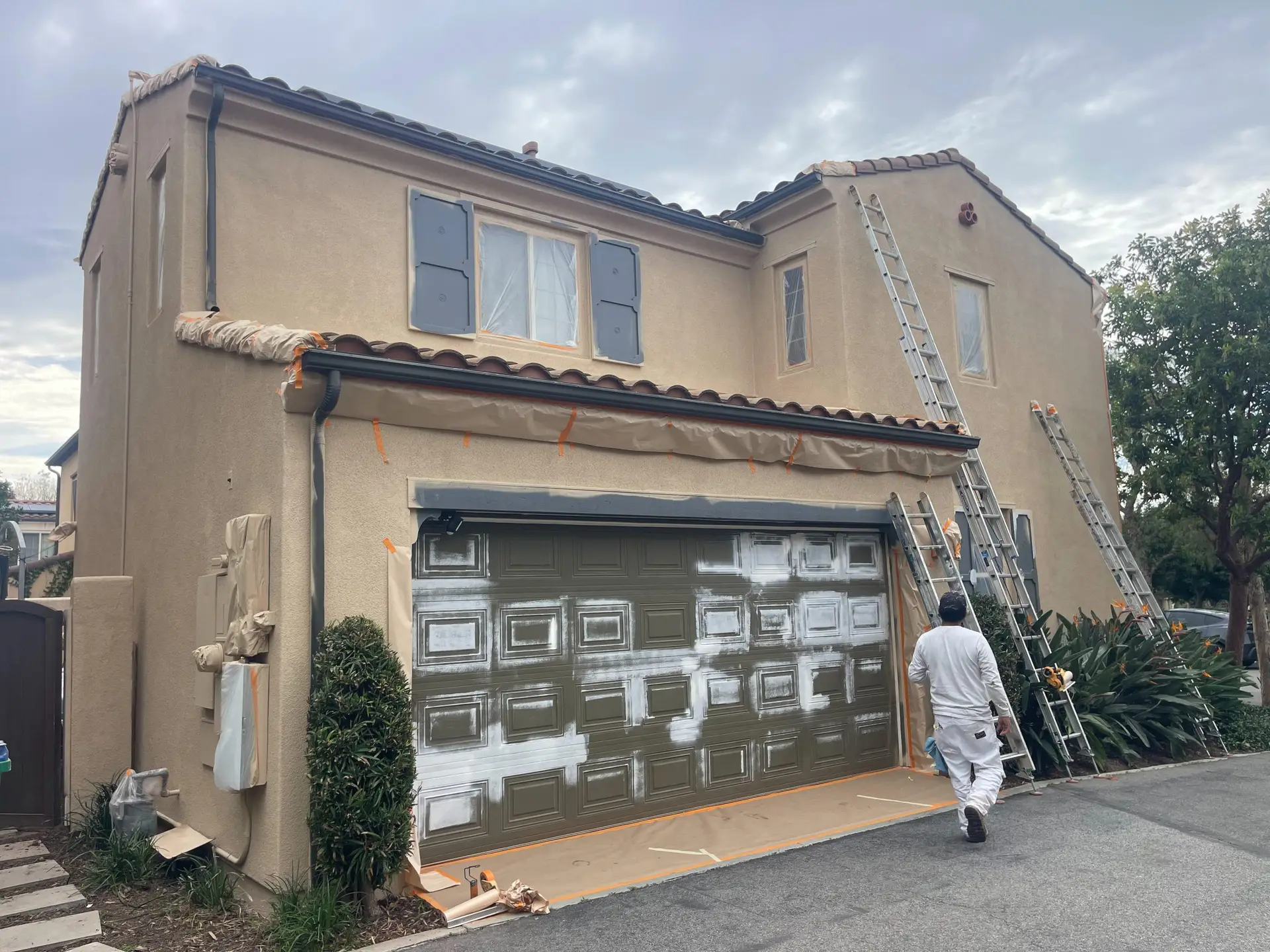 Beige two-story house under renovation with ladders against the facade and a garage door being painted, orange tape securing protective coverings, cloudy sky.