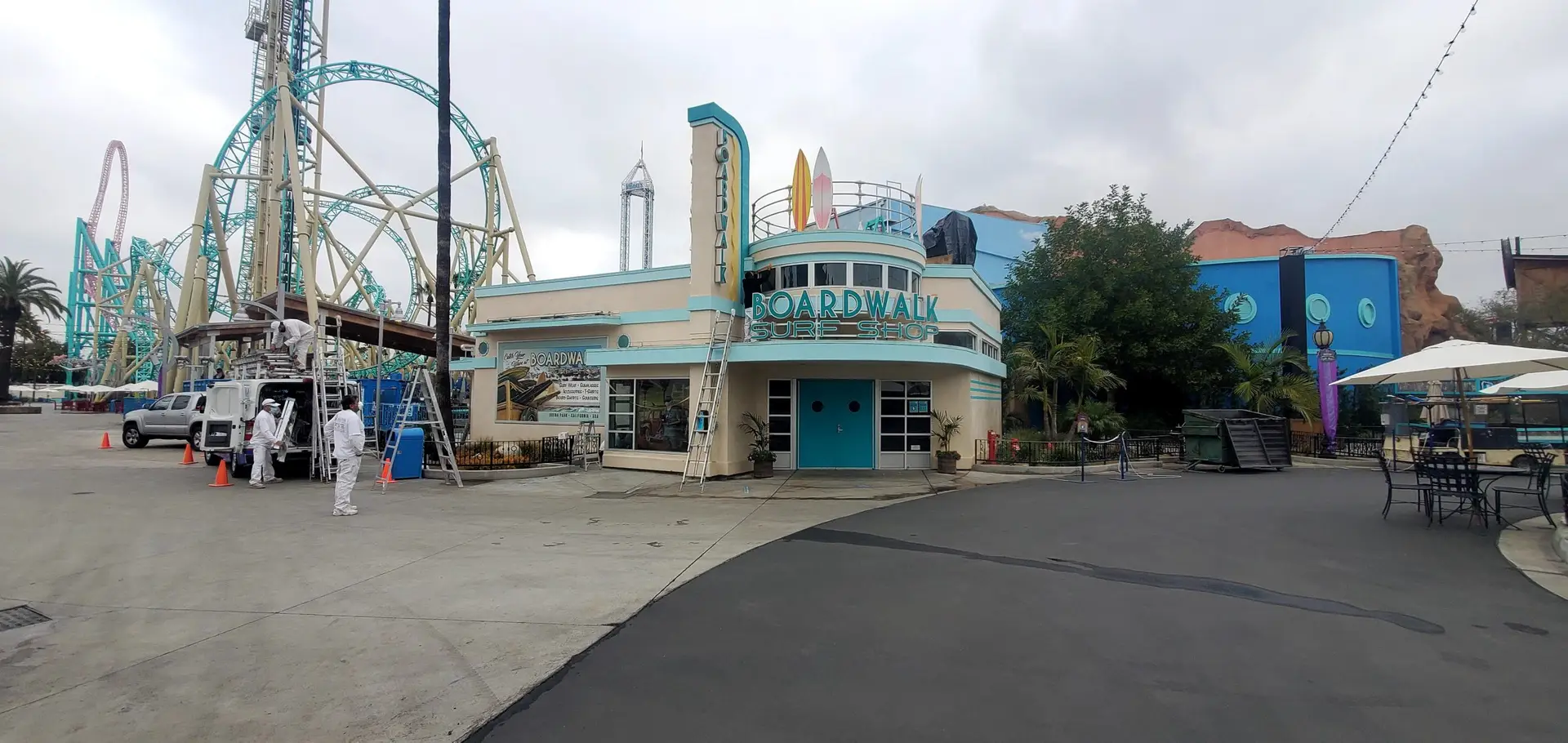 Amusement park scene with a retro-style Surf Shop building labeled 'Boardwalk Surf Shop' and a large turquoise roller coaster in the background, workers in white uniforms nearby.