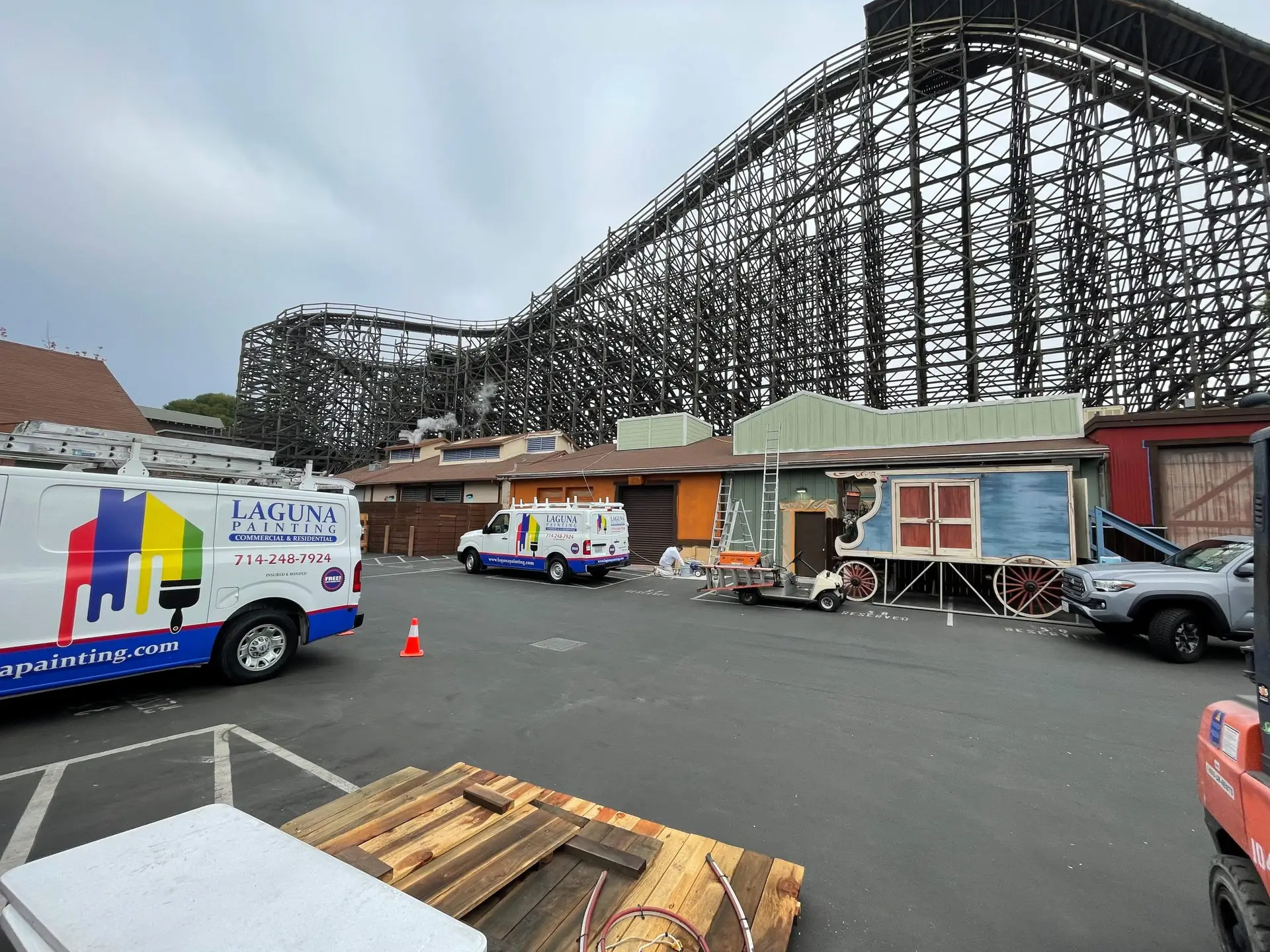 Construction area around a massive wooden roller coaster frame, with service vans and a decorative wagon in the foreground