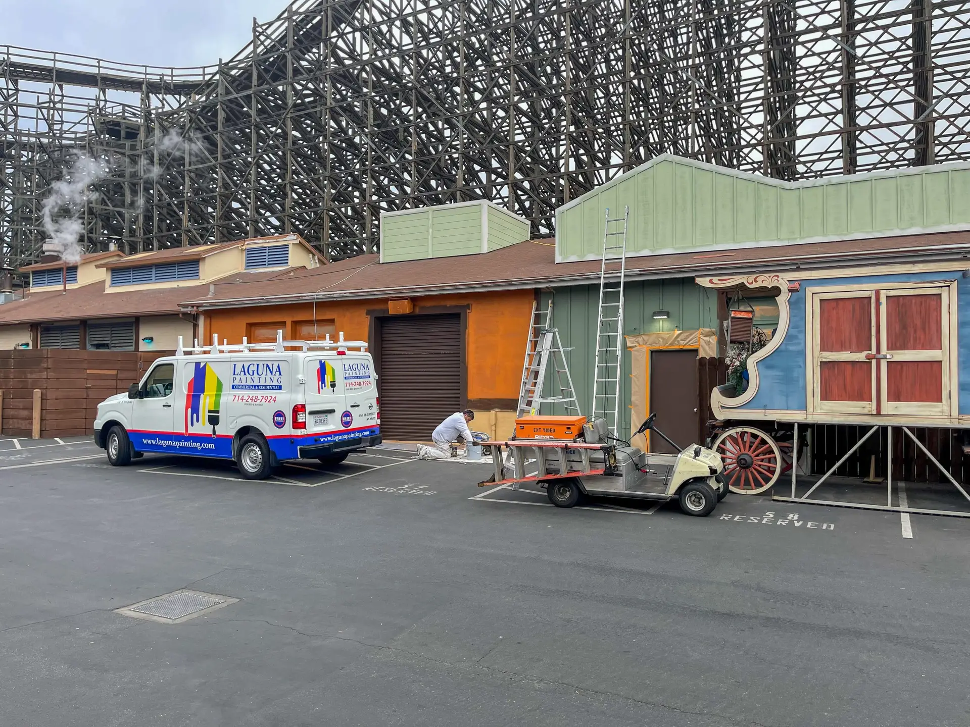 Painting crew with ladders and tools beside a decorative wagon in a parking lot; a Laguna Painting van is parked nearby with a wooden roller coaster frame in the background.