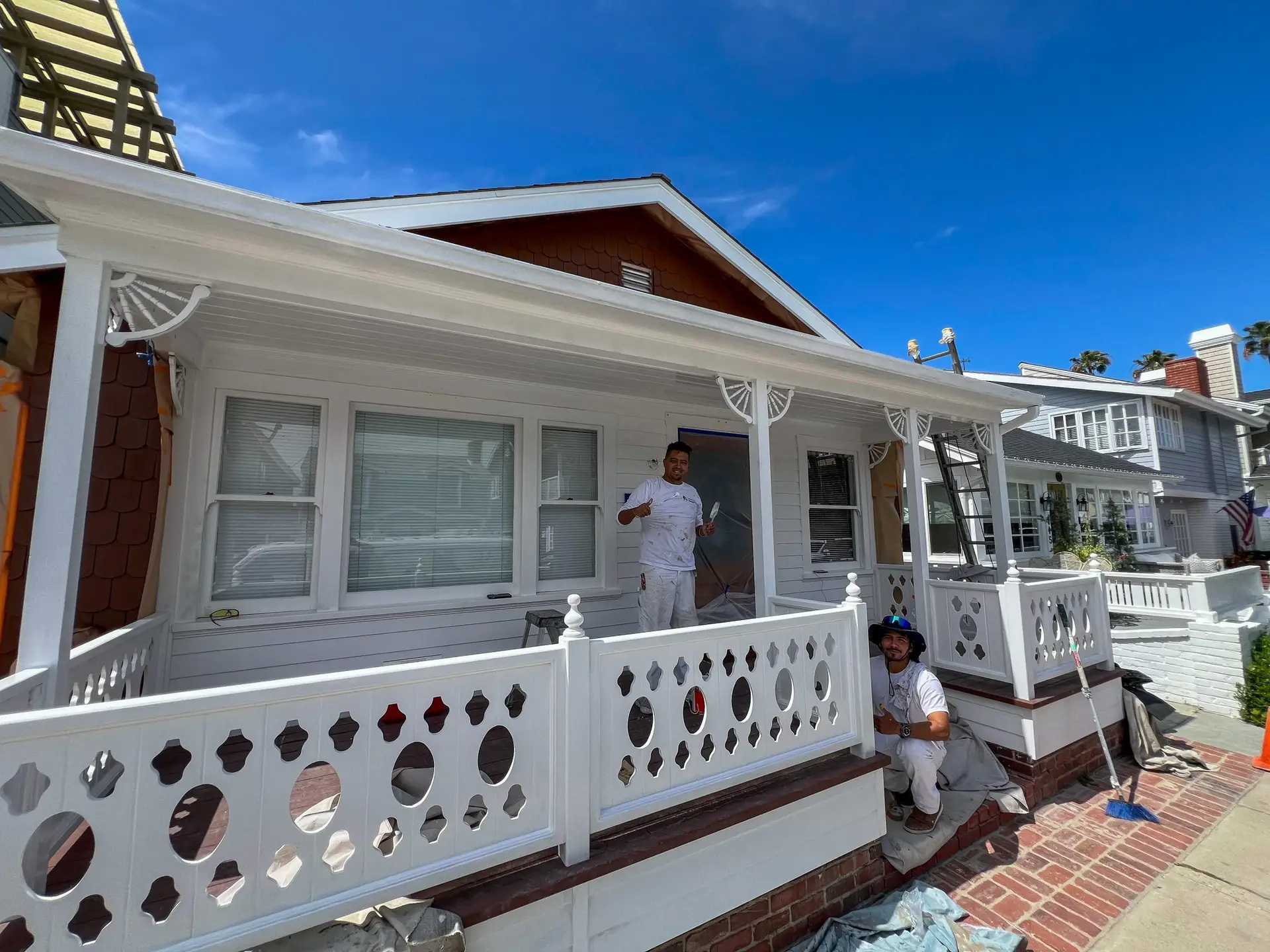 Two painters work on a white front porch with decorative cutout railing on a sunny day