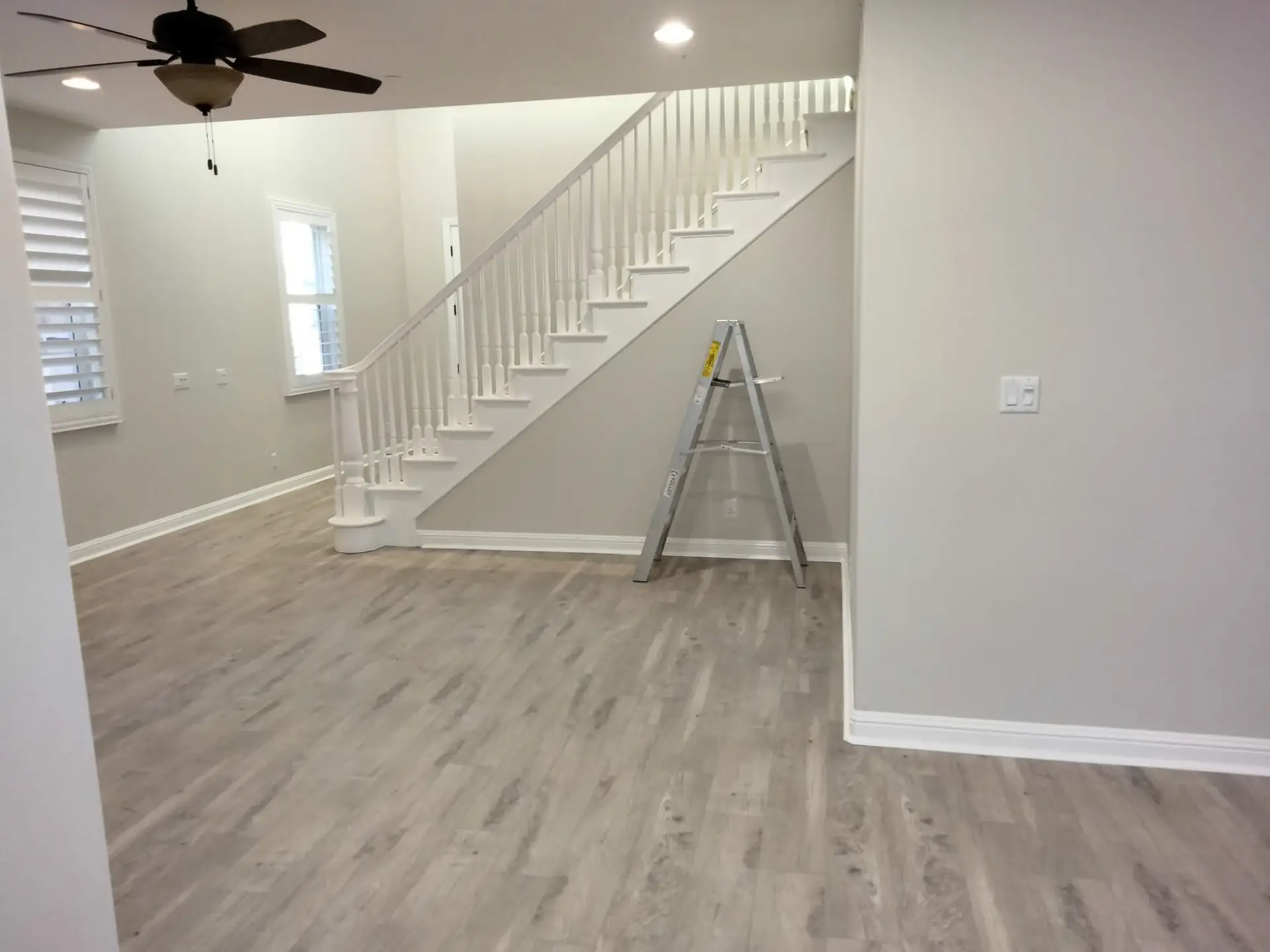 Interior living area with gray walls and light wood flooring; white staircase with balusters along diagonal wall, and a simple ladder set against it, indicating renovation in progress.
