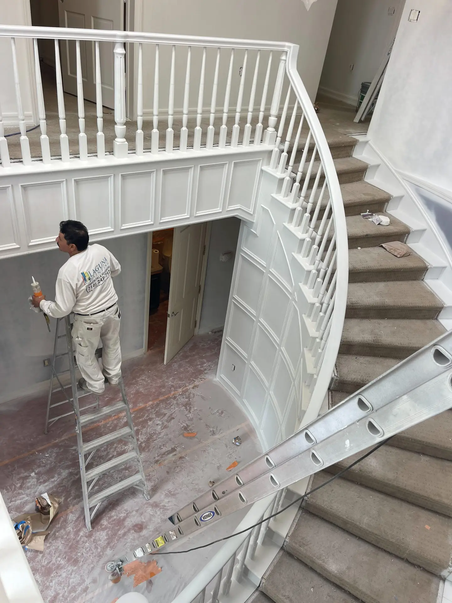 A worker on a ladder painting a white curved staircase with balusters and wainscoting in a home under renovation.