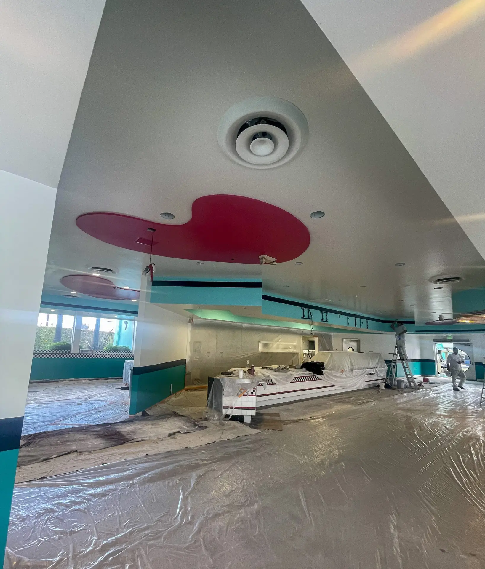 Under-construction retro diner interior with turquoise walls, pink ceiling shapes, and plastic-covered floor as workers paint.