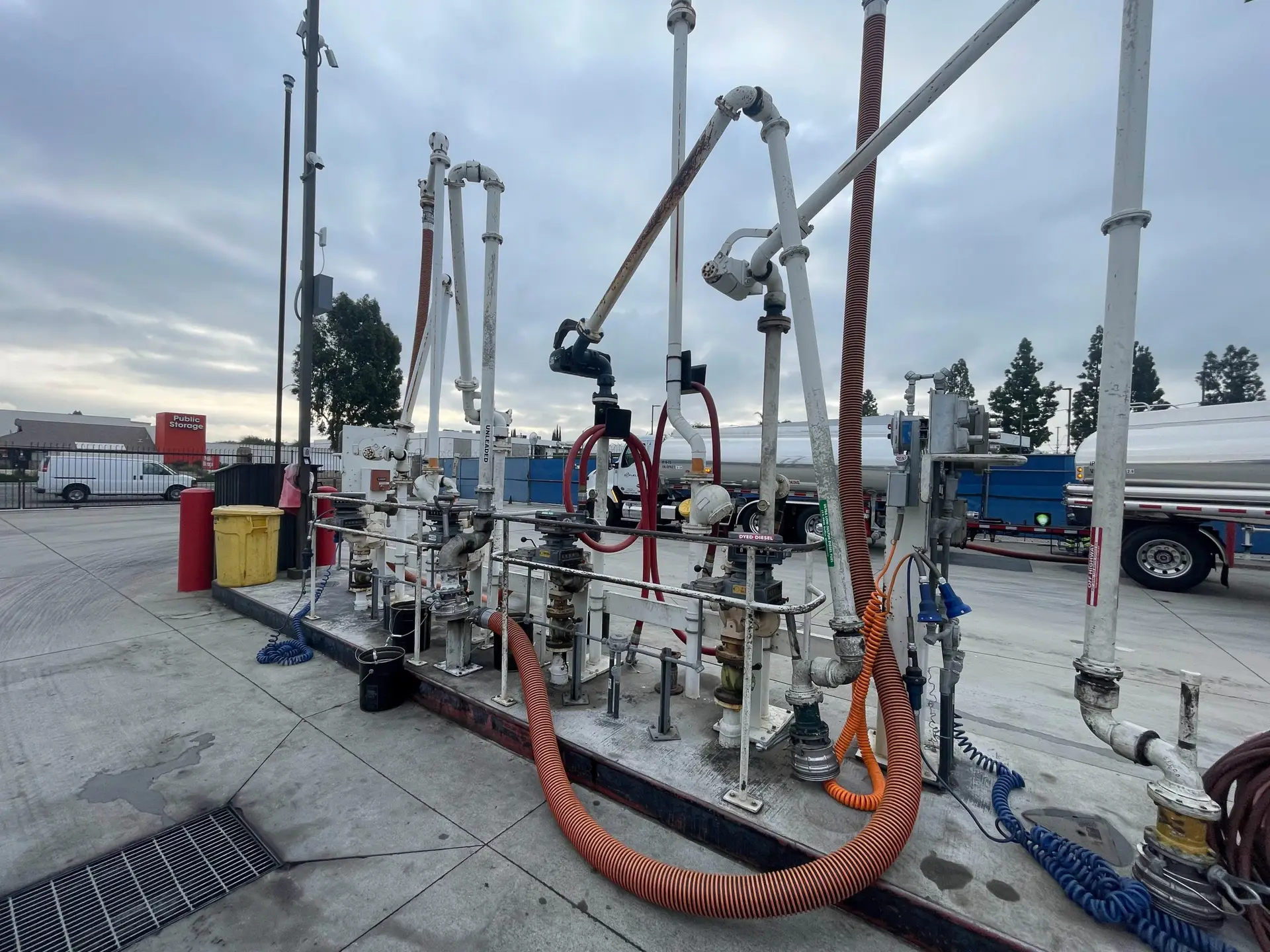 Industrial fueling rack with multiple pipes, hoses, and valves at a concrete station, likely for diesel or gas transfer. A large orange corrugated hose and blue coiled hose are visible, along with metal frames and gauges. Overcast sky in the background and a yellow trash bin nearby.