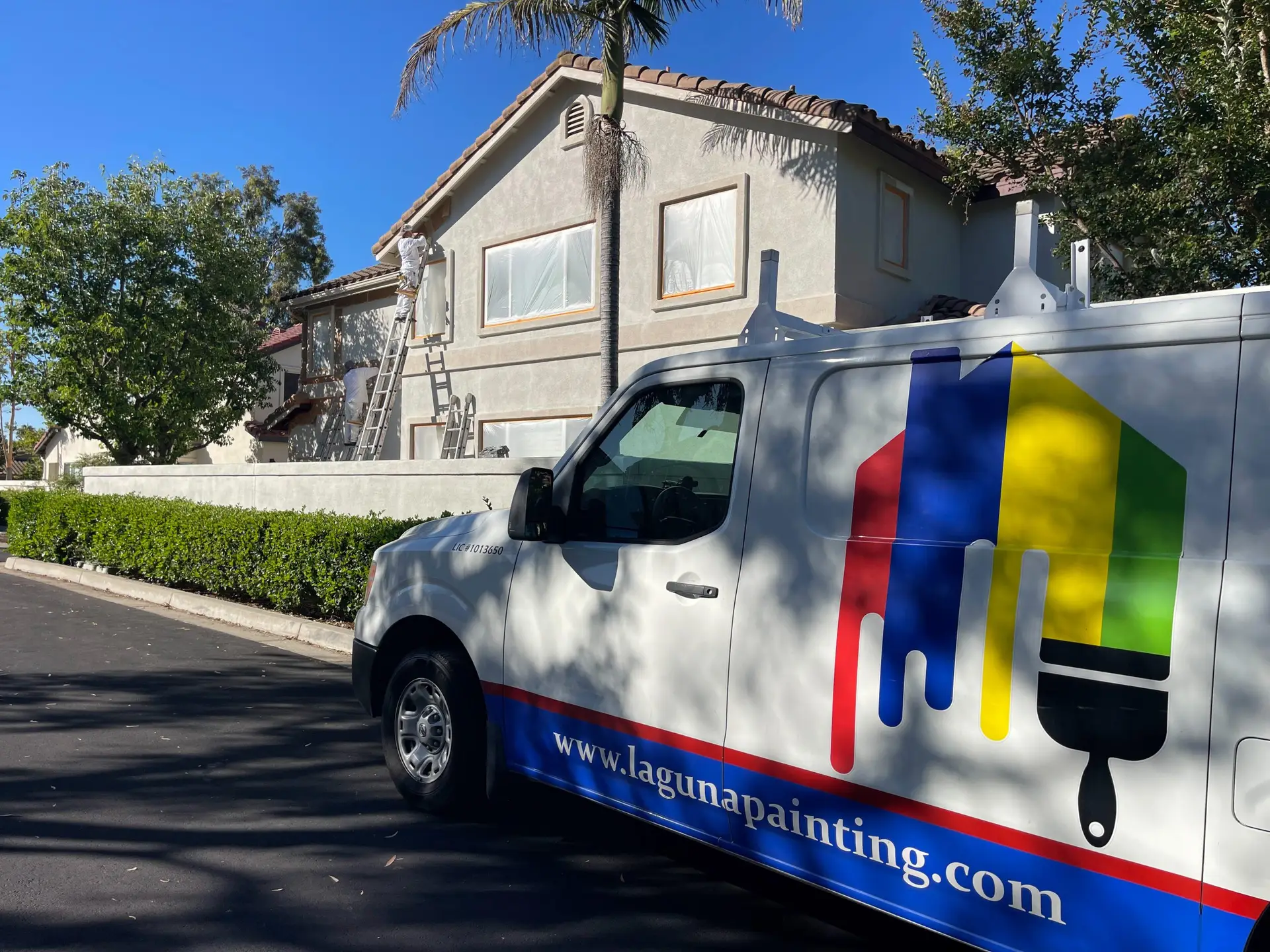 Three painters work on ladders to paint the beige two-story house, with a colorfully logoed painting van in the foreground.