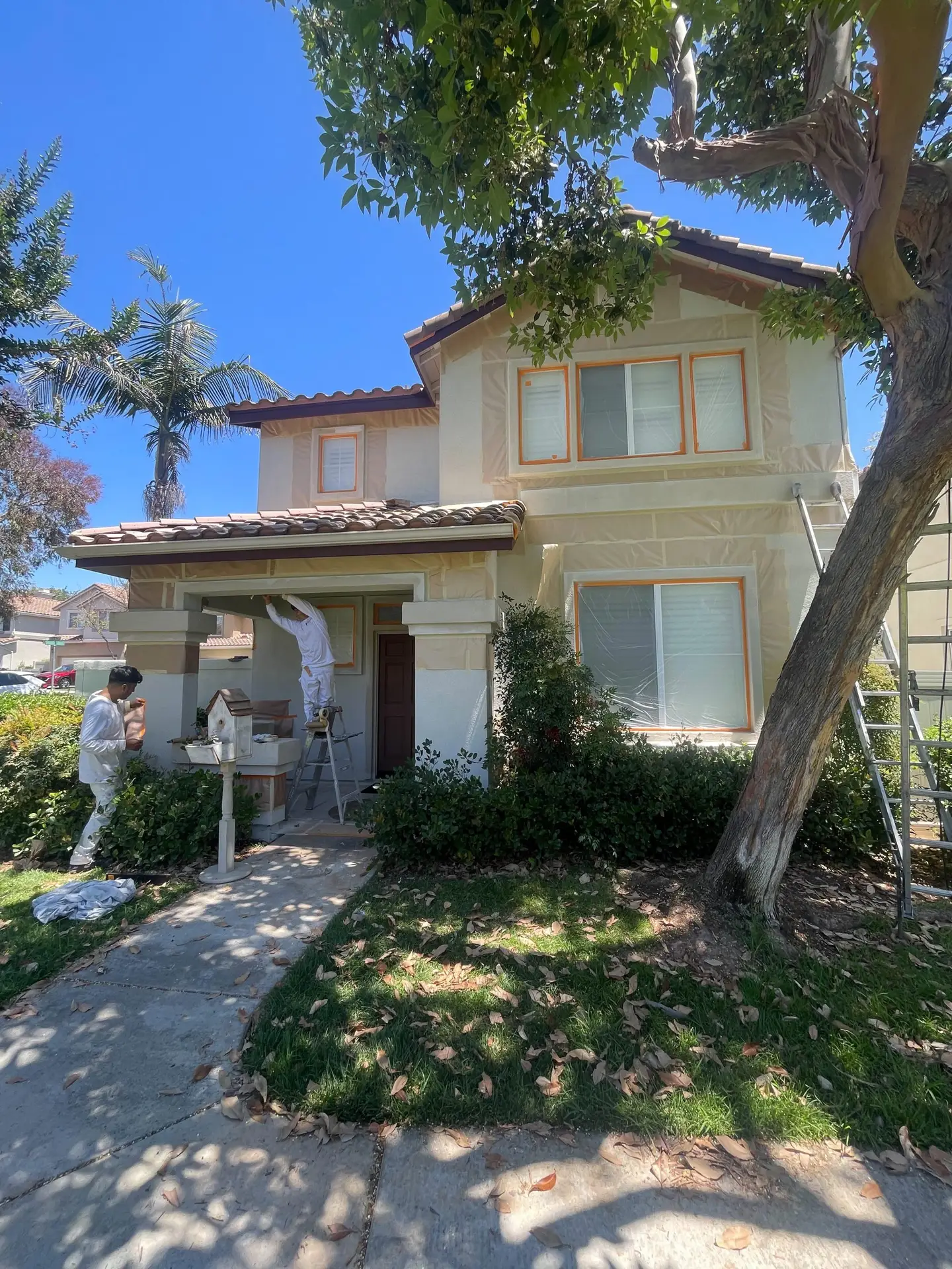 Two workers in white coveralls painting the beige two-story house, with a ladder at the front and plastic sheeting around windows awaiting finishing touches.
