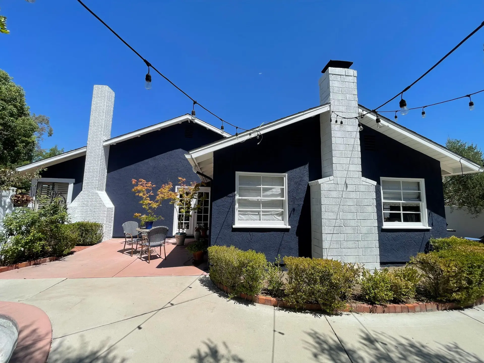Blue stucco house with two white chimneys, string lights overhead, and a peach-colored patio with a small table and chairs.