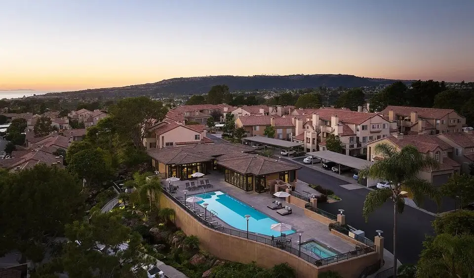 Aerial view of a suburban apartment complex with a rectangular blue pool, poolside seating, and a clubhouse at sunset, surrounded by beige townhouses.
