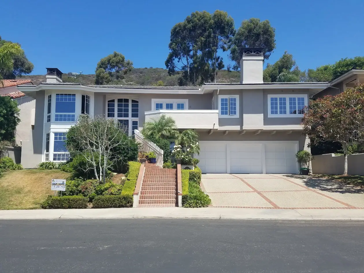 Two-story gray suburban home with large white-framed windows, brick stairway leading to the front door, and a three-car garage; hillside and trees in the background. A small real estate sign is in the front yard.