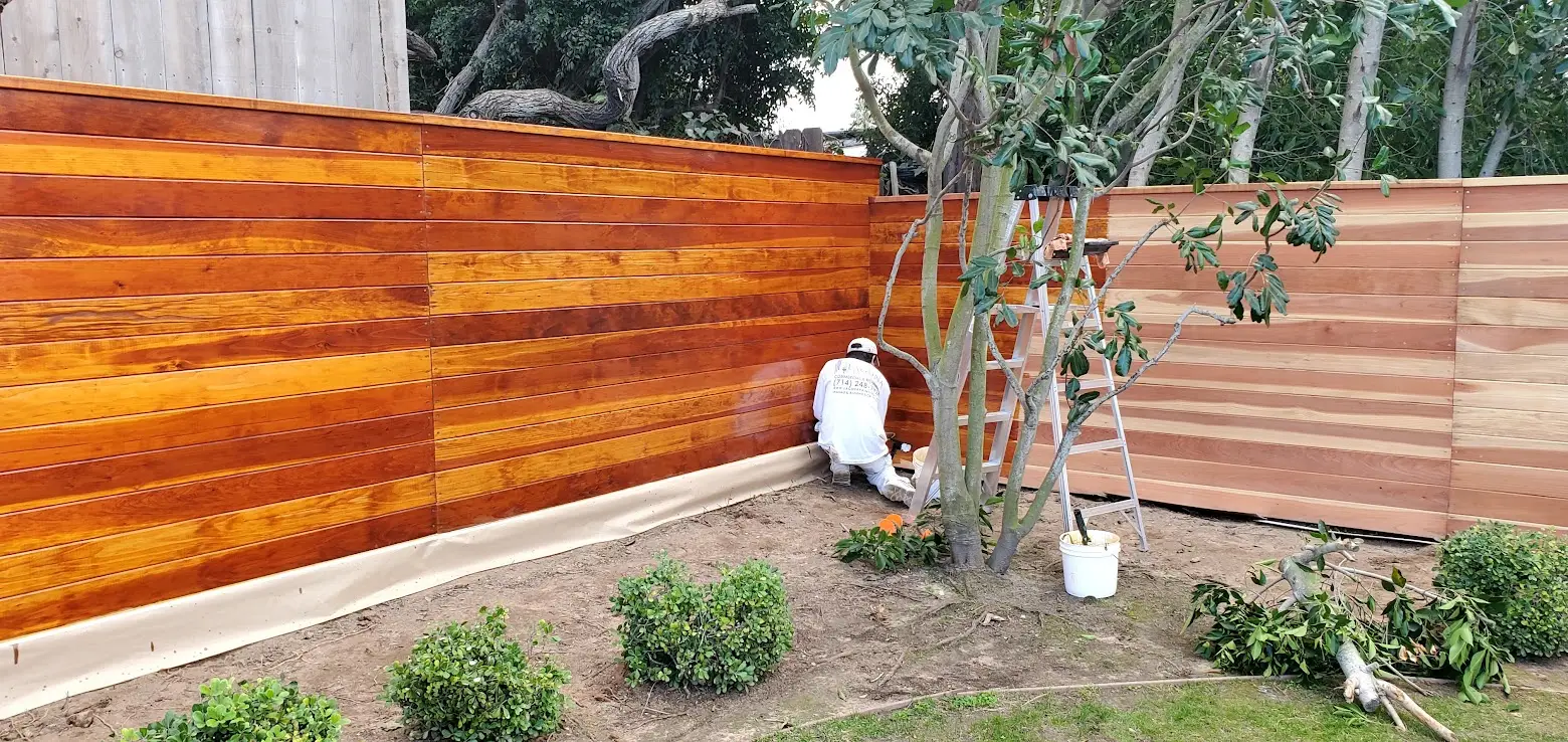 Worker kneeling by a tall, freshly stained wooden fence in a backyard, with a ladder and bucket nearby.