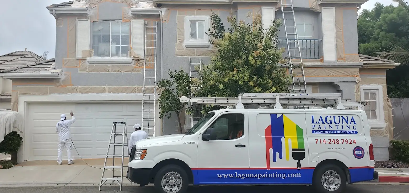 Exterior painting in progress on a gray two‑story house; workers in white overalls with ladders and a Laguna Painting van parked out front.