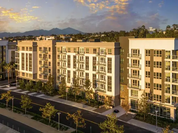 Modern apartment complex with beige buildings, green landscaping, and a mountain range in the distance at sunset in the background.”