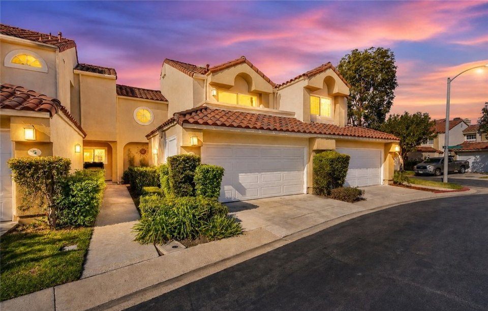 Row of beige stucco townhouses with tiled red roofs, three garages, and well-kept shrubs under a sunset sky.