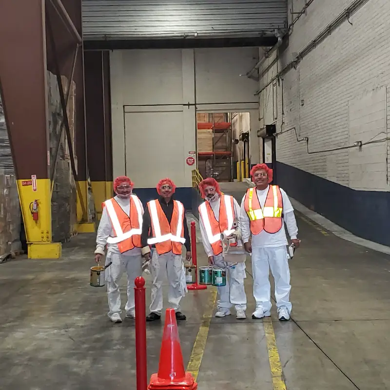 Group of five workers in orange safety vests and hair nets standing in a warehouse, holding paint cans.