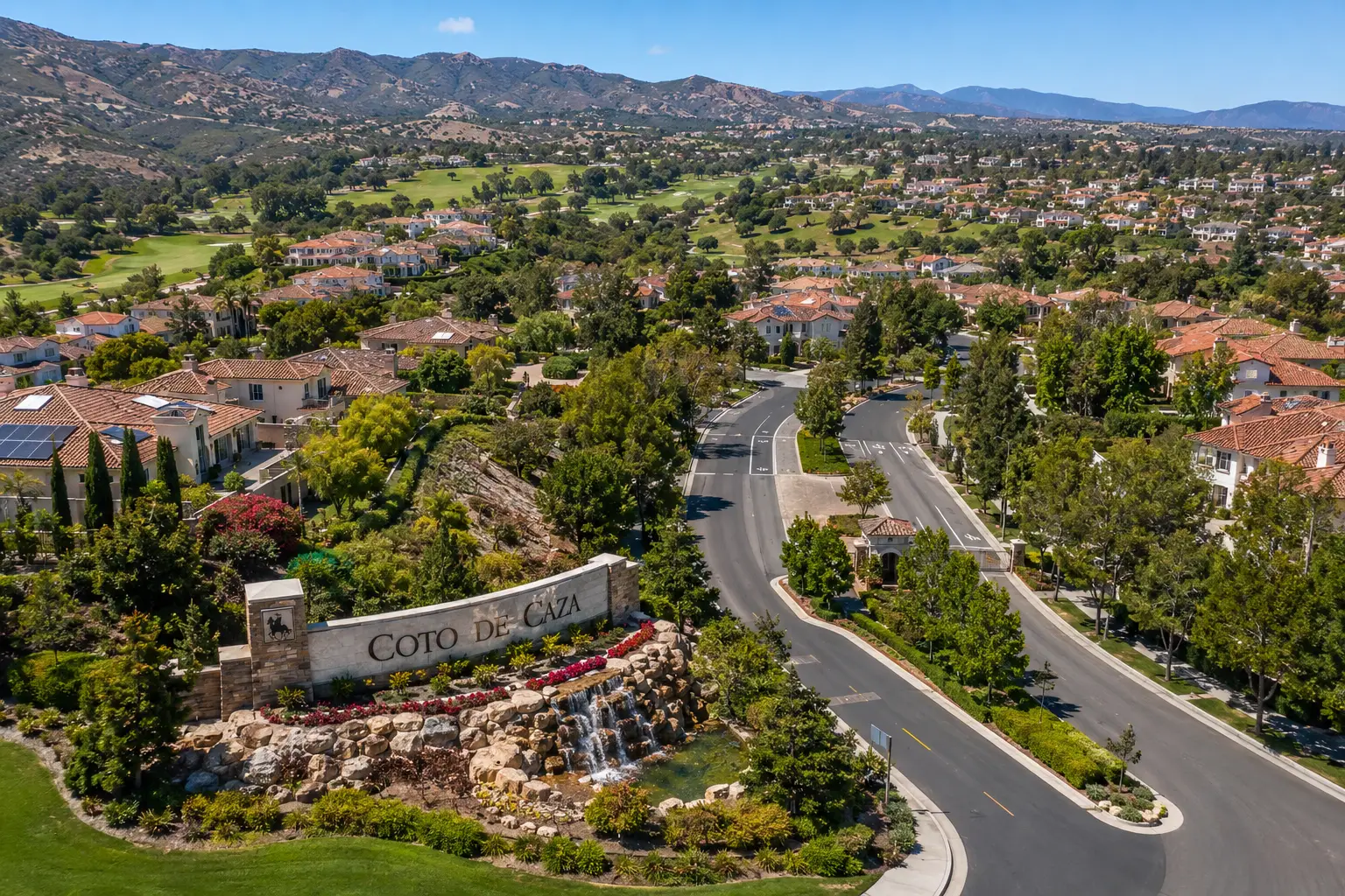 Aerial view of the Coto de Caza entrance with a stone sign and cascading rocks, surrounding with lush landscaping and curved roads.