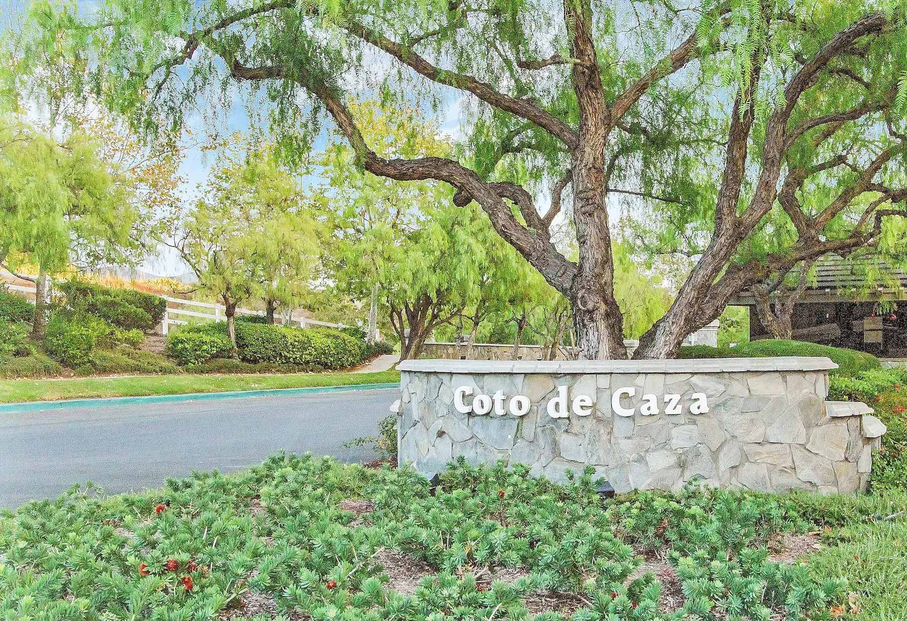 Stone sign reading 'Coto de Caza' built from gray stone, surrounded by shrubs and trees in a landscaped area at a road entrance.