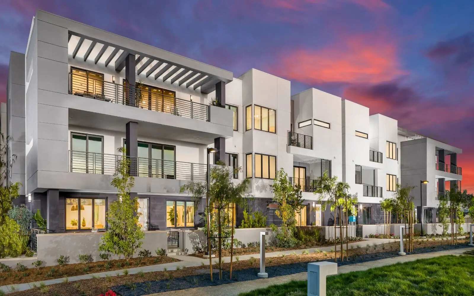 Modern white apartment complex with balconies and landscaping at dusk, glowing interior lights.