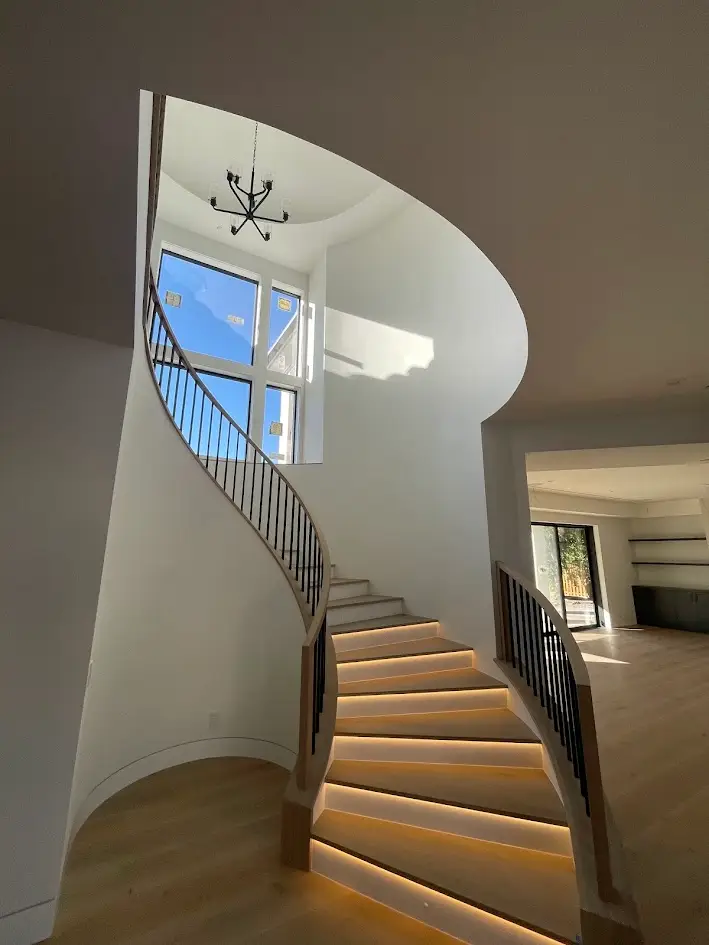 Spiral wooden staircase with metal balusters curves up a bright white atrium, topped by a modern chandelier and large windows.