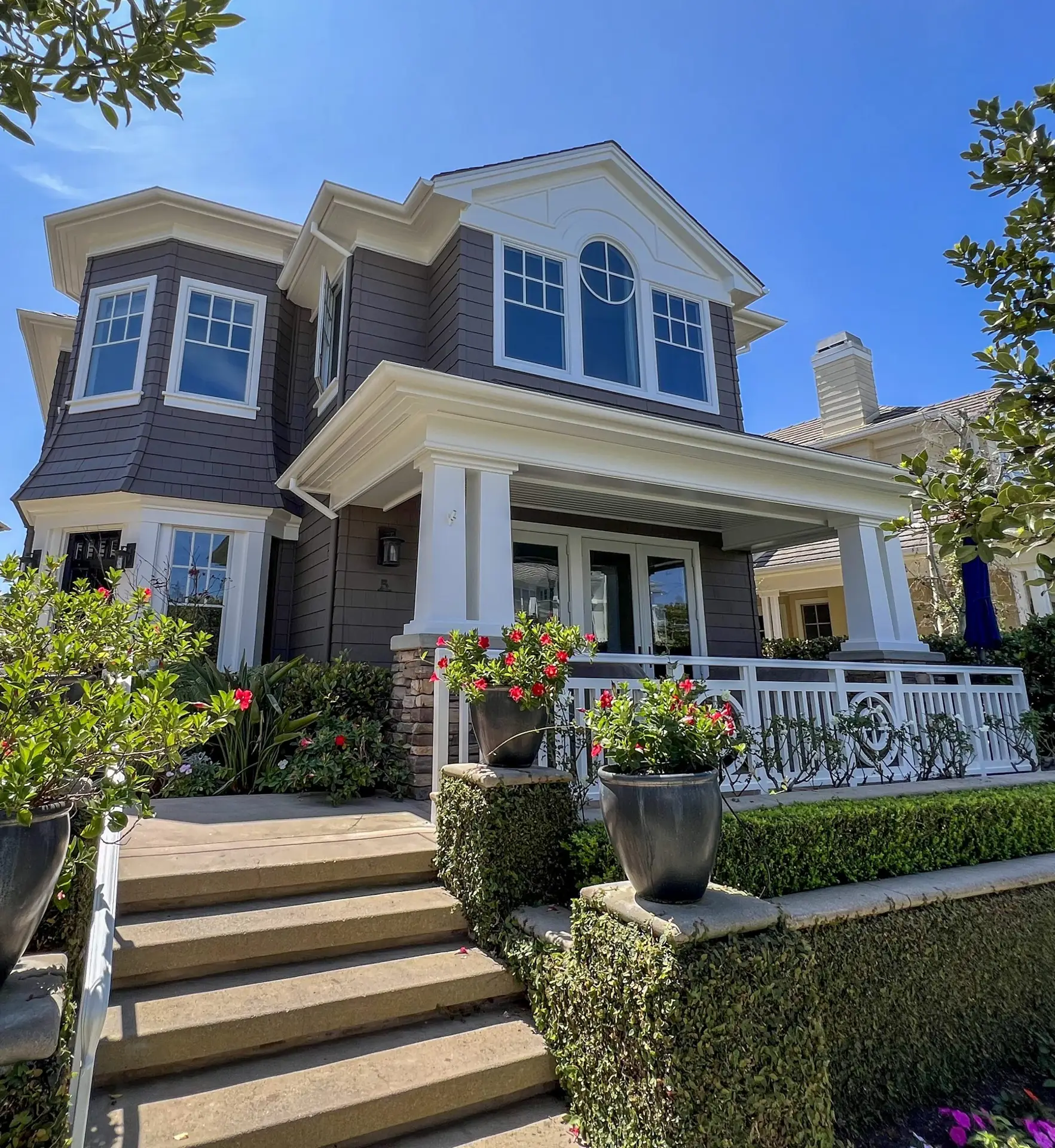 Two-story house with brown siding, white trim, and a prominent arched window above the entry, surrounded by plants and a white fence.