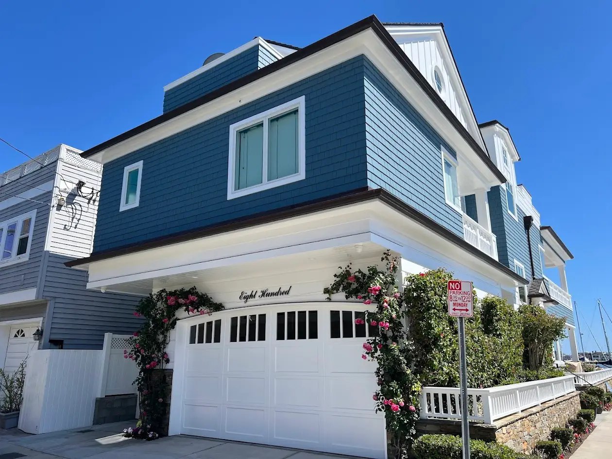 Blue coastal house with white trim and a large white garage door, roses climbing along the entrance, under a clear blue sky.