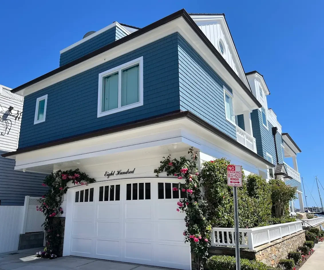 Blue seaside-style house with white trim, a large white garage door, and pink climbing roses against a clear blue sky.