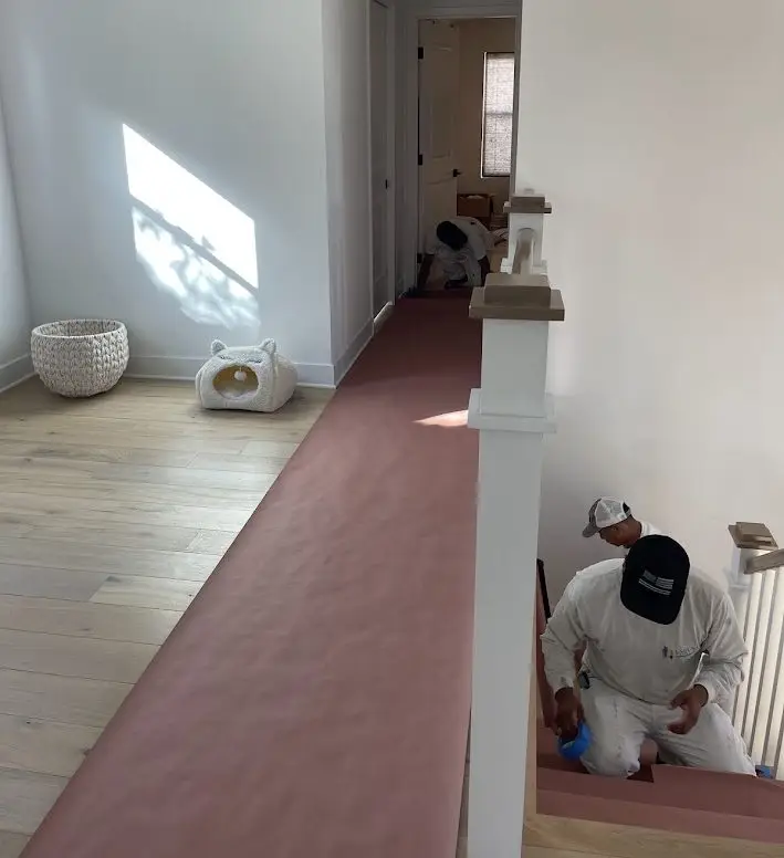 Bright upstairs hallway under renovation with a pink carpet runner; a worker in white overalls descends the stairs.