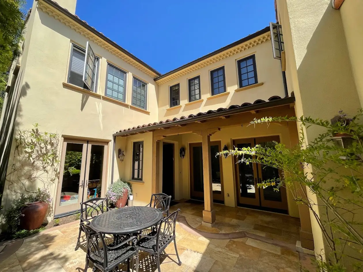 Sunlit Mediterranean courtyard with beige stucco walls, iron patio table and chairs, and potted plants by glass doors and windows.