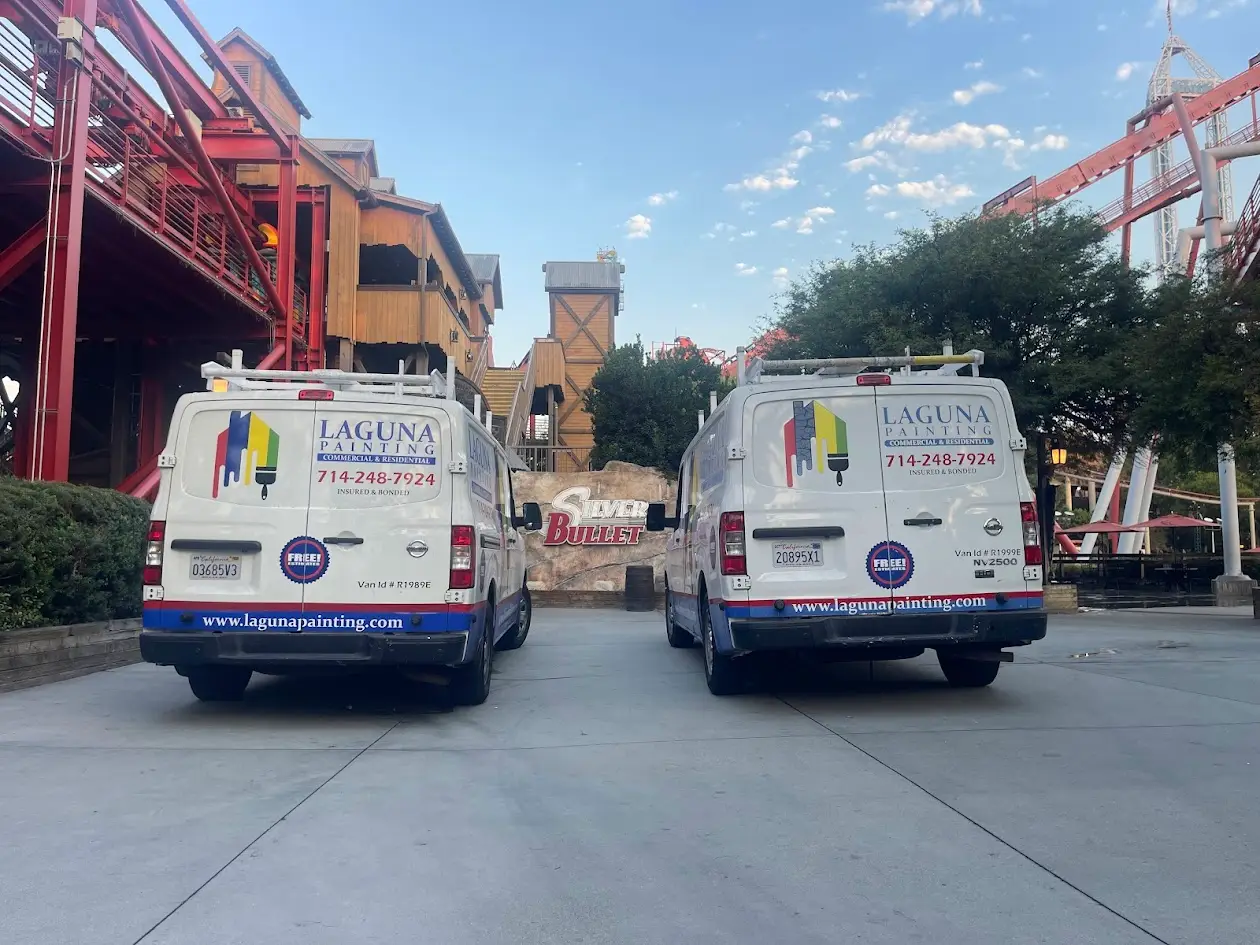 Two white Laguna Painting service vans parked side by side in front of a wooden theme-park building with red steel framework and a clear blue sky behind.