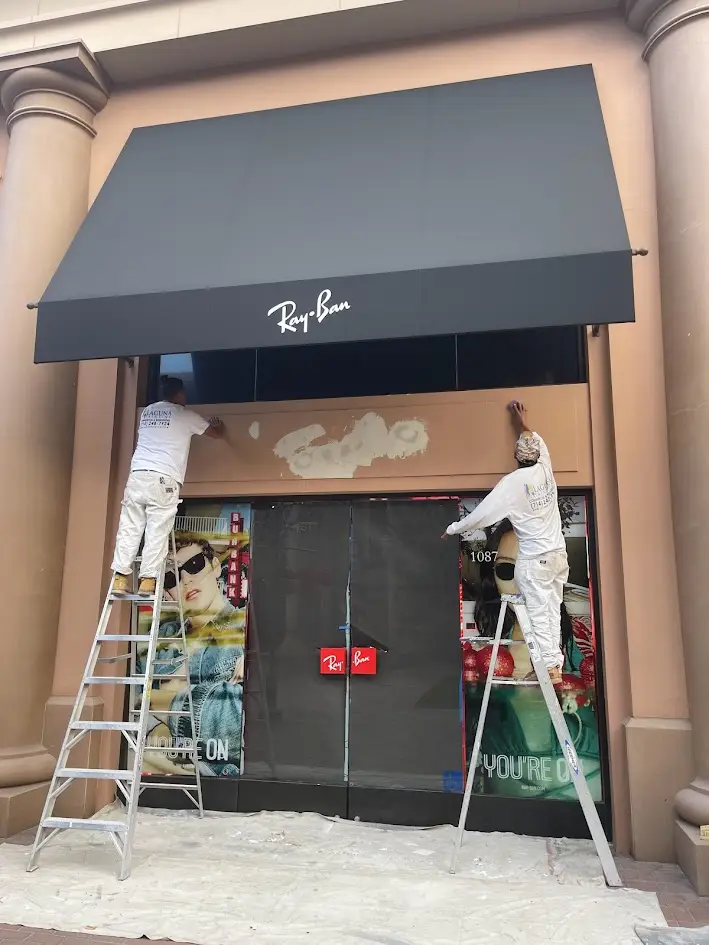 Two painters on ladders repair the beige storefront beneath a large black Ray-Ban awning.