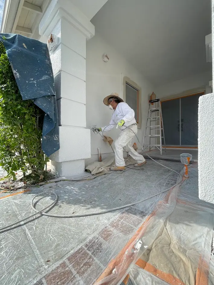 Painter in protective gear spraying a white column on a covered porch, with a ladder nearby and plastic sheeting on the ground