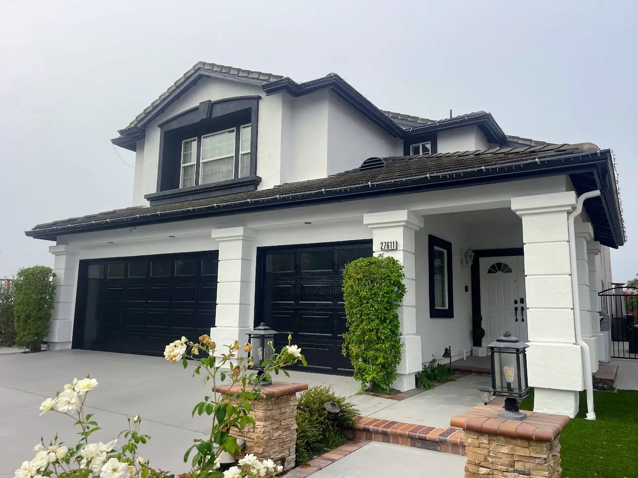 Two-story white house with black trim and a double garage, front porch, and landscaped walkway with roses in bloom.