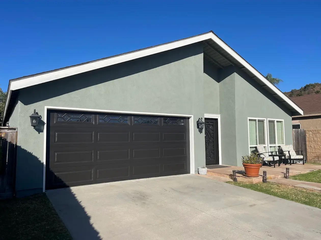 Green-gray suburban house with a dark sectional garage door, front entry door, and a small seating area on the right with chairs and a potted plant outside.