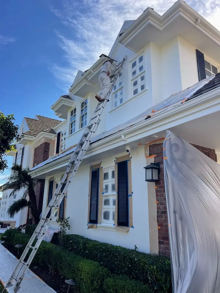 A person on a tall ladder painting the white exterior of a two-story house with dark blue shutters under a blue sky.