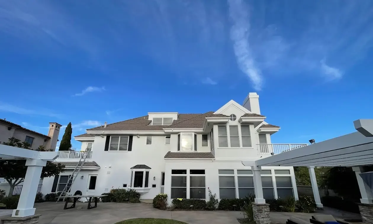 Backyard of a white two-story house with large windows and a gray roof, a curved lawn in the foreground, and a white pergola against a clear blue sky.