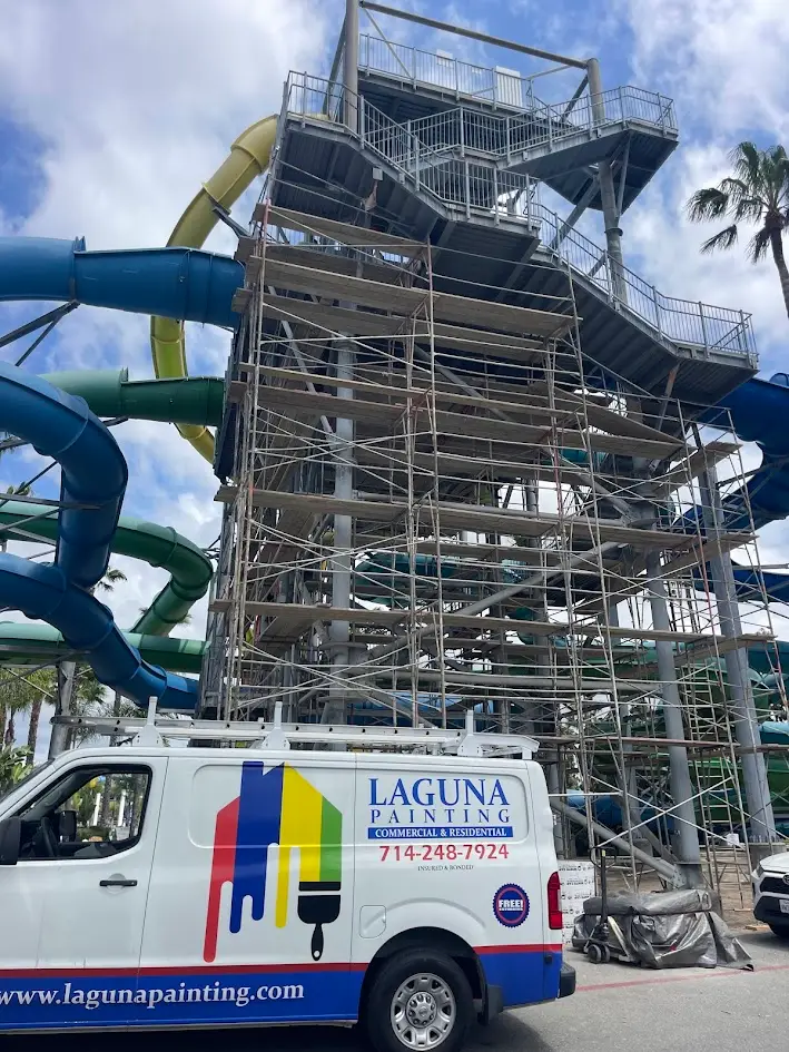 Tall water slide tower under construction with extensive scaffolding and colorful slide tubes, a Laguna Painting van parked in front.