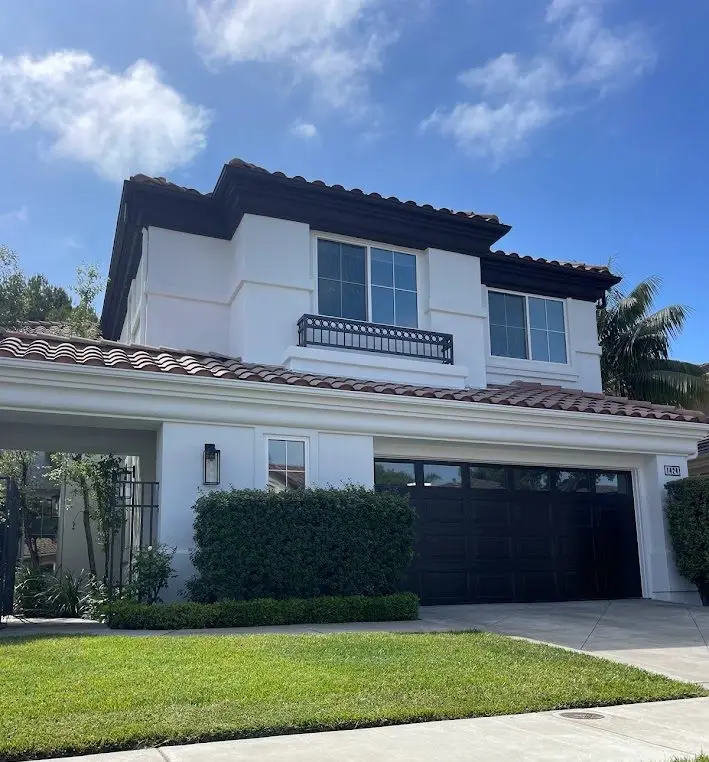 Front view of a two-story white stucco house with a red tile roof and a black garage door, manicured shrubs, and a blue sky.