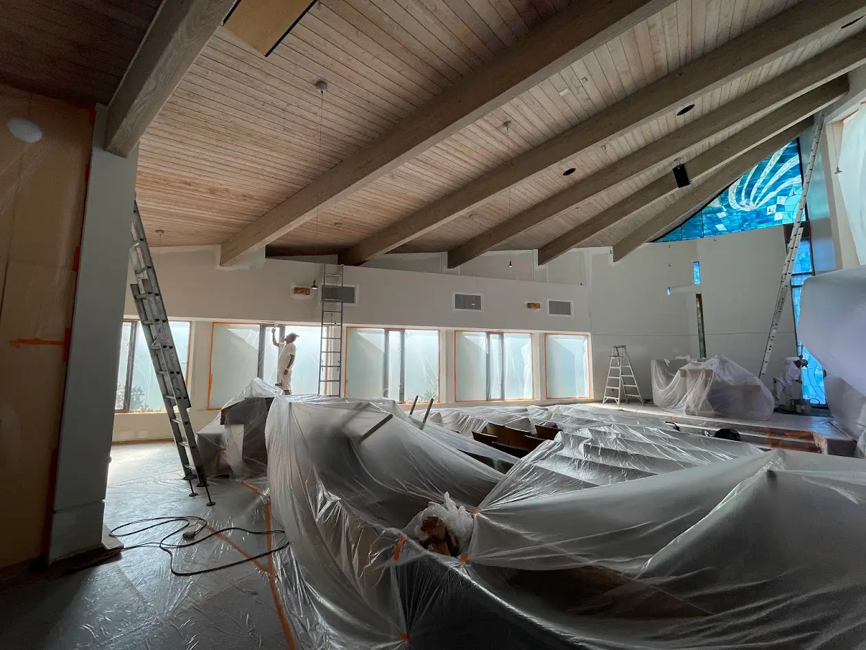 Large indoor construction site with workers, plastic-covered equipment, and tall wooden ceiling beams. Ladders leaning against walls as renovation continues.