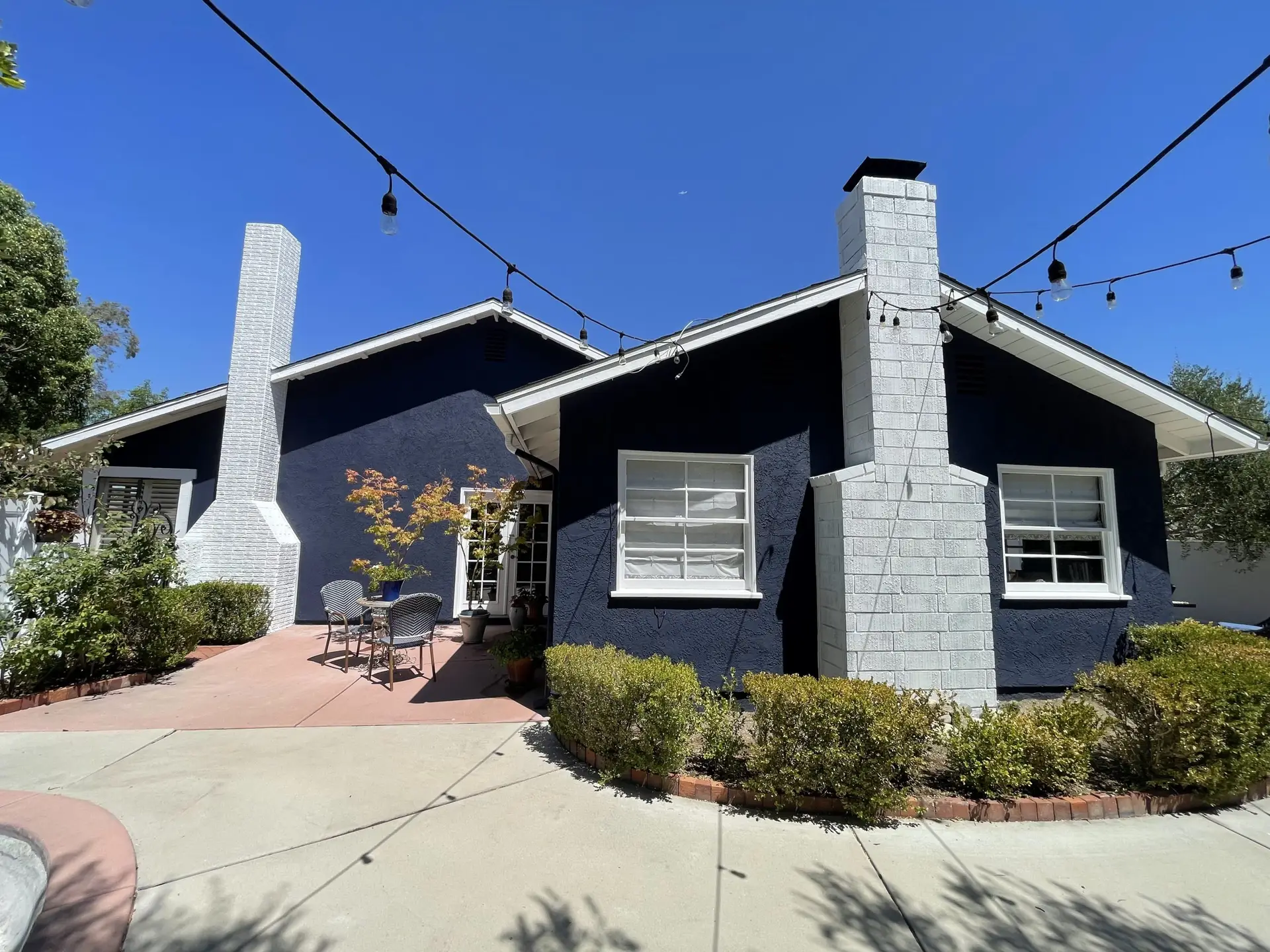 Front-yard patio area of a navy house with white brick chimneys and string lights overhead under a clear blue sky.