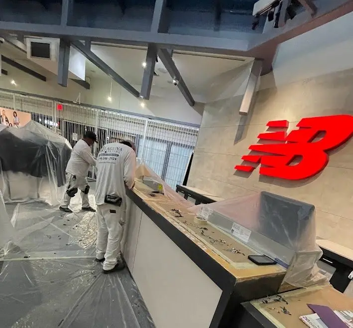 Interior of a retail store under construction with workers in white coveralls and plastic sheeting on the floor and counters, large red New Balance logo on the wall.