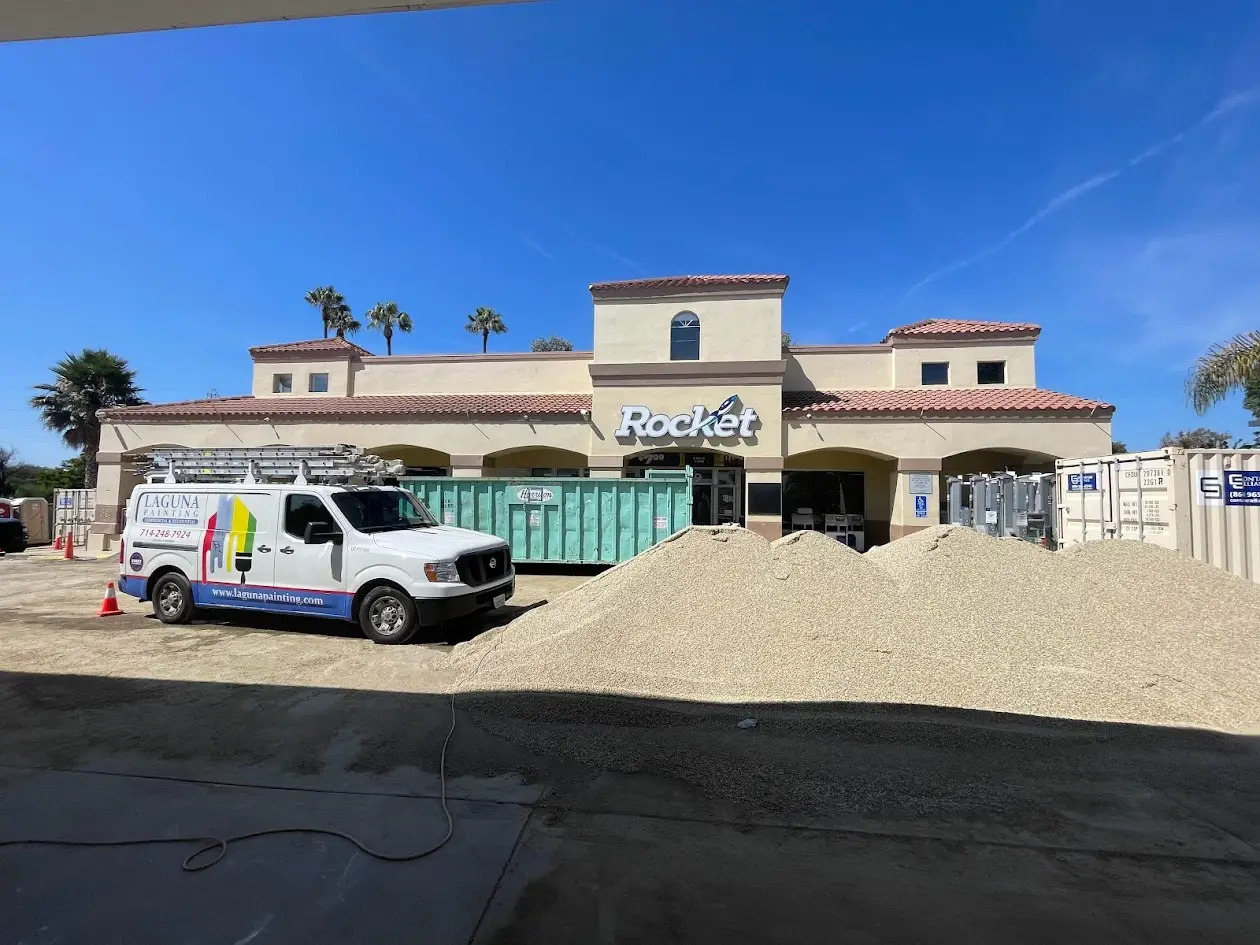 Rocket store undergoing renovation with a large gravel pile in front and a Laguna Painting van nearby for scale and work scaffolding under a clear blue sky.