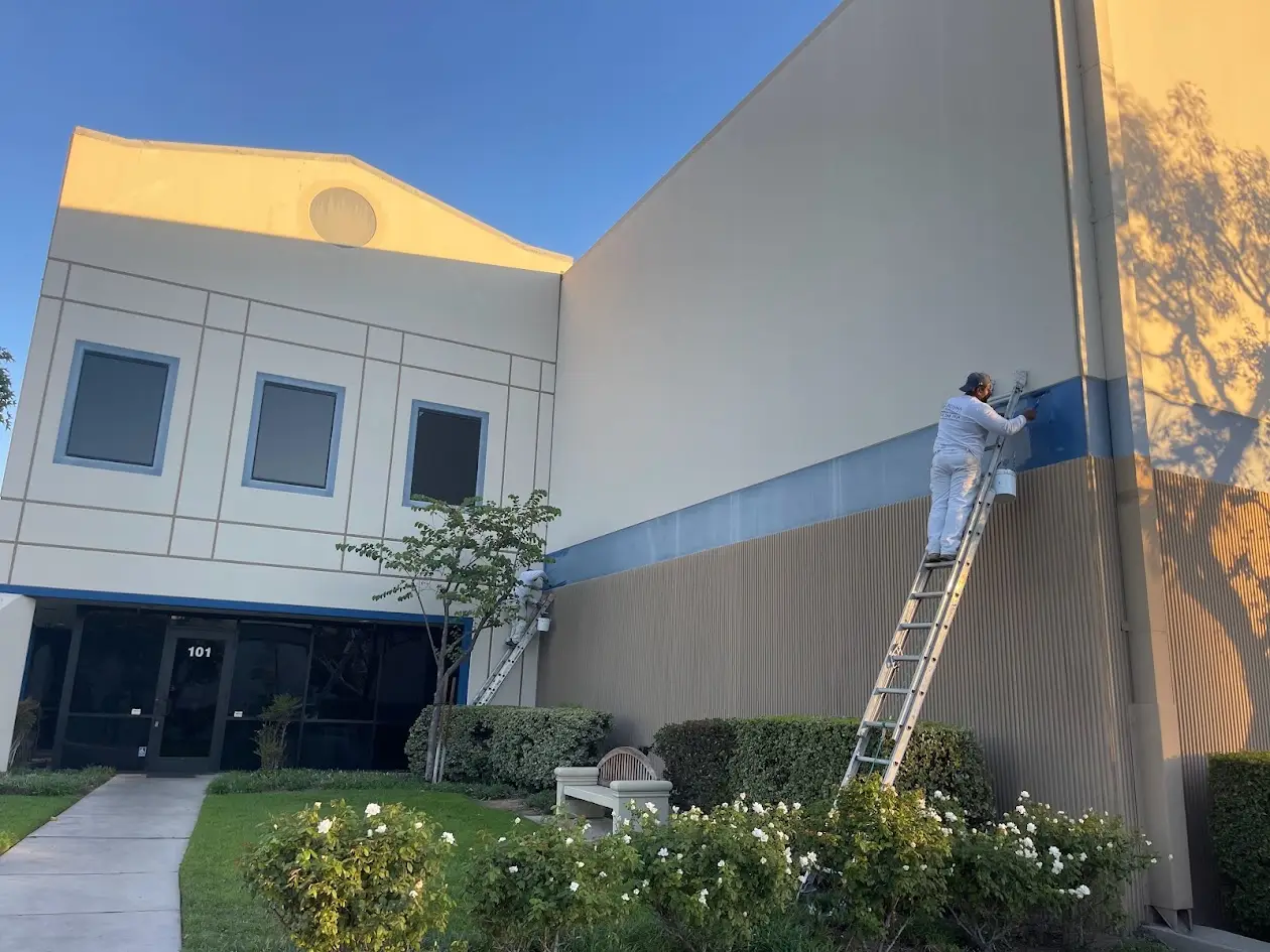 Two workers painting a blue stripe on the beige exterior of a commercial building; one on a ladder applying paint, with a bucket nearby.