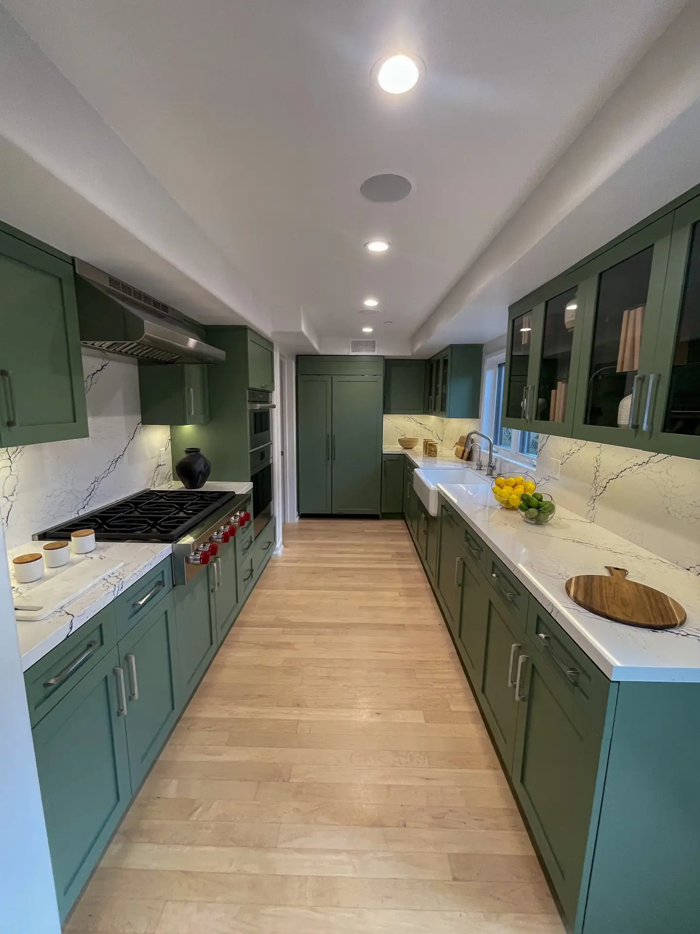 Long, narrow kitchen with dark green cabinets, marble countertops, and a row of ceiling lights along both walls.