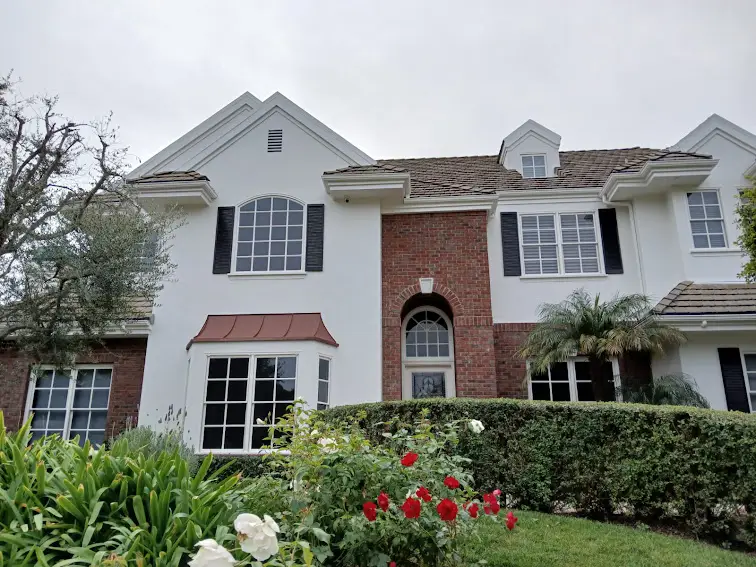 Front view of a two-story white and brick house with an arched brick entryway and black shutters, surrounded by a manicured lawn and rose bushes.